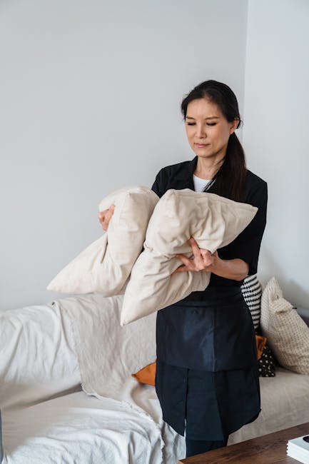 A person wearing a black jacket is performing surface cleaning on a mustard yellow cushion with a white geometric pattern, placed on a dark fabric sofa in a room with plain white walls. The individual